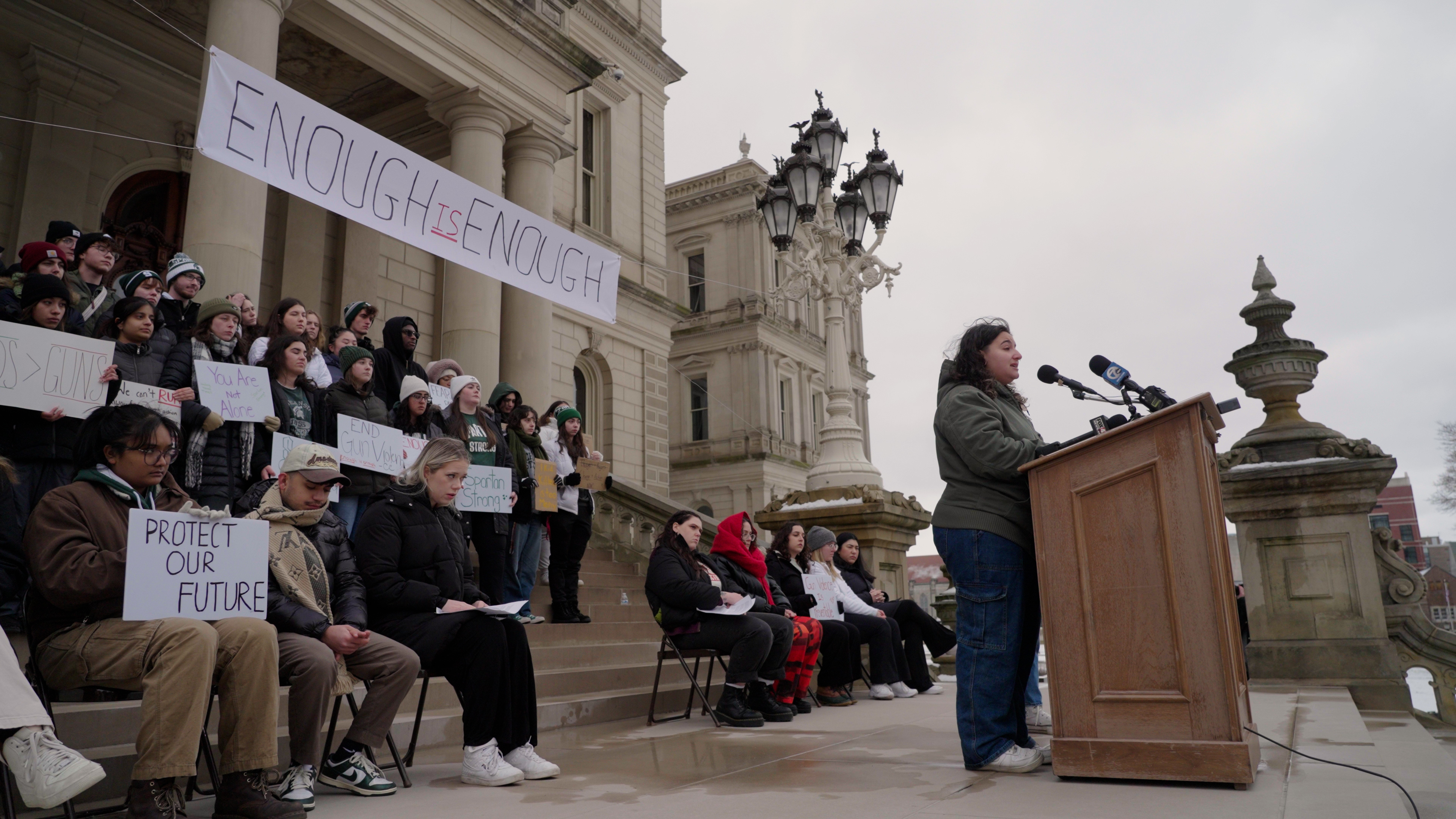 A woman standing at a podium in front of a protest at the Michigan capitol building