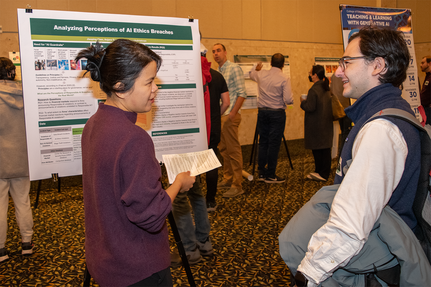 Two people talking in front of a research poster, with other people in the background.