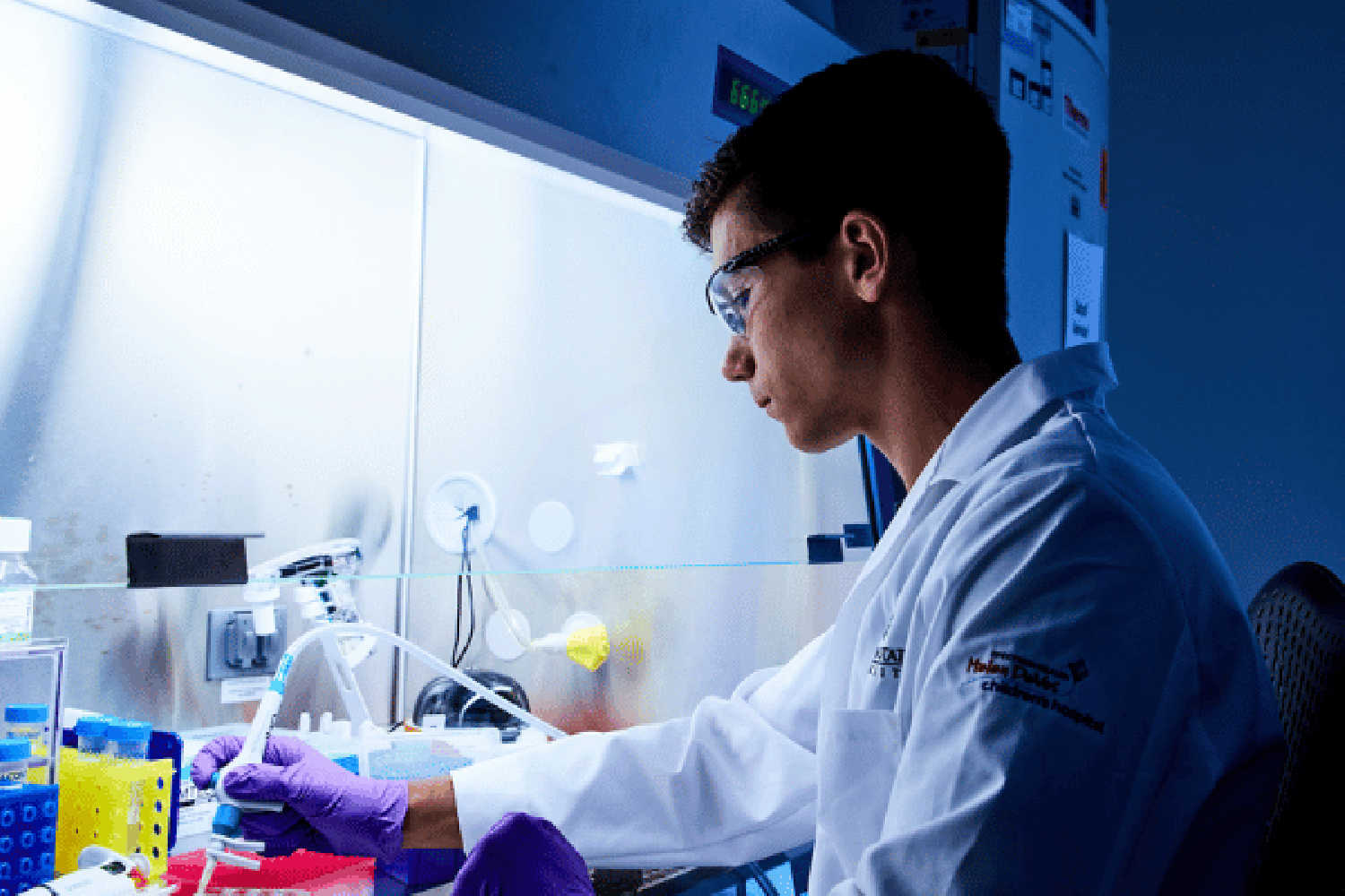 A person in a white lab coat and safety glasses working in a lab. 