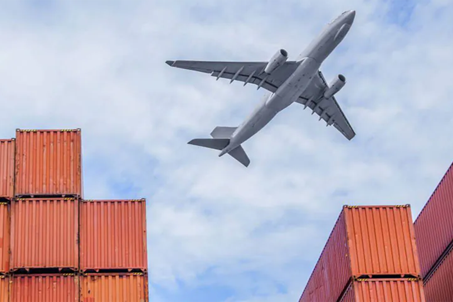 A large airplane flying over stacked orange shipping containers. 