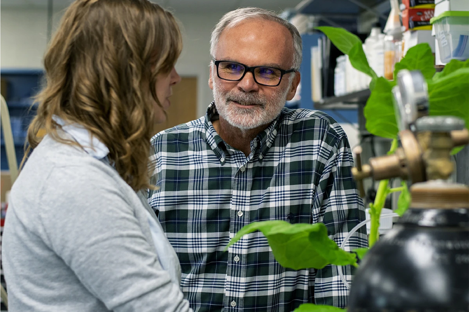 A professor in a plaid shirt and glasses talks to a student in a white lab coat. 