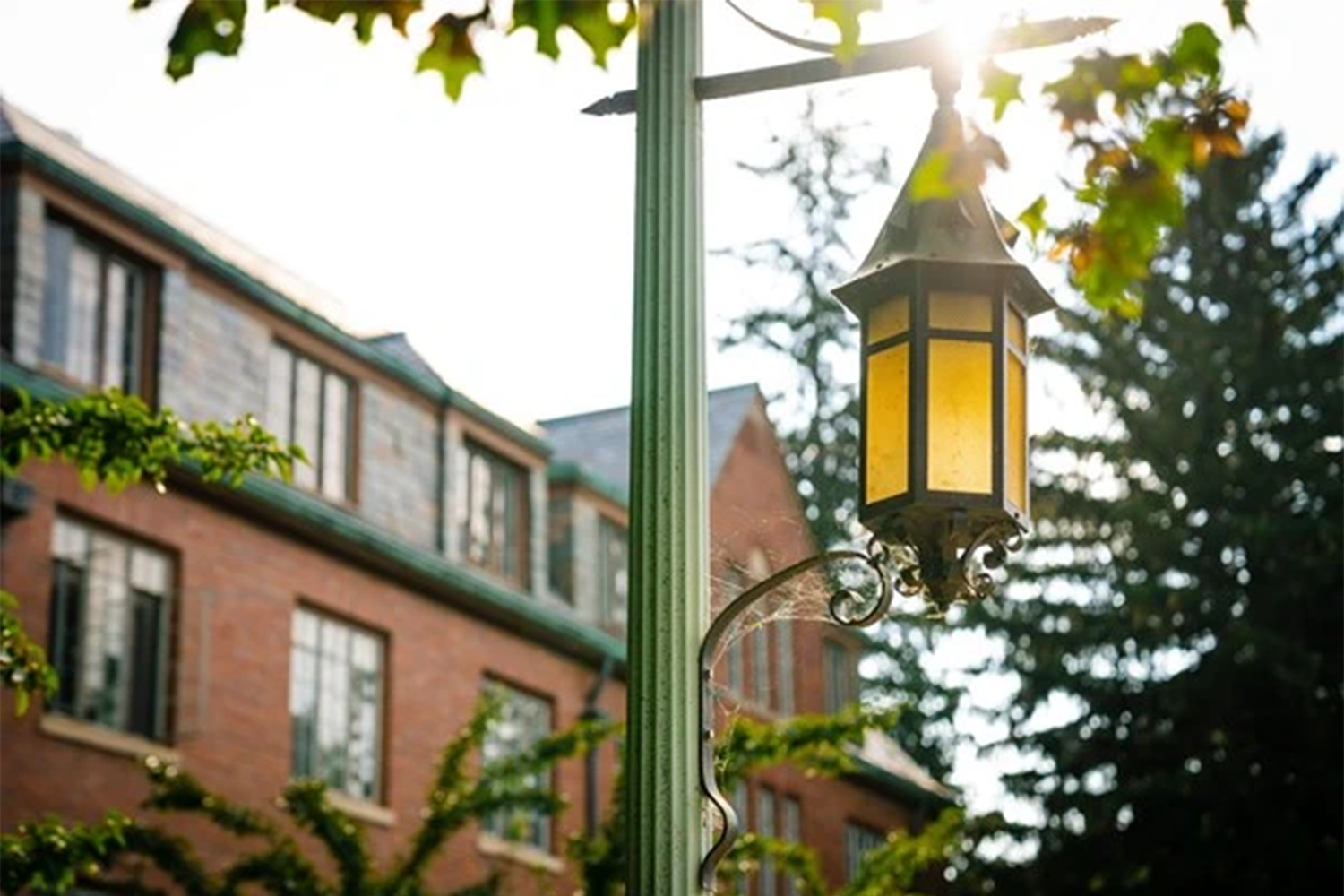 A lantern on a post with a red brick building and trees in the background.