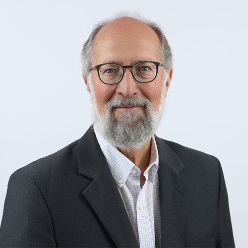 A man with grey hair and beard, glasses, wearing a light shirt and dark jacket, in front of a white background