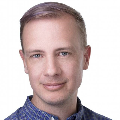 A man with short blonde hair, and a shirt with a blue collar