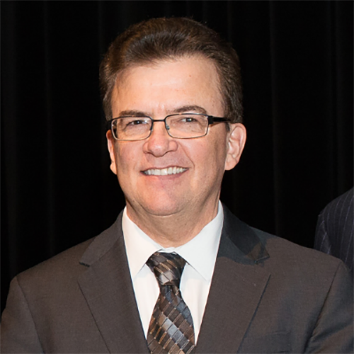 A man with short dark brown hair, glasses, wearing a brown suit and tie in front of a black background