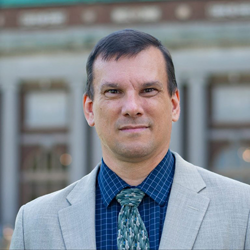 A man with short dark hair, a blue collared shirt and tie, and a light suit jacket, standing in front of a building