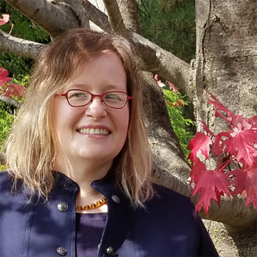 A woman with mid-length blonde hair and red glasses, wearing a dark blue jacket and top, with a tree and leaves behind her