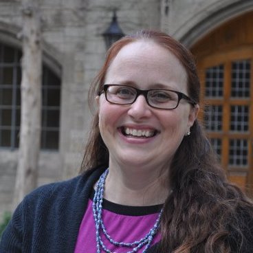 A woman with long brown hair and glasses, wearing a long blue necklace, purple shirt, and dark jacket, with a building in the background