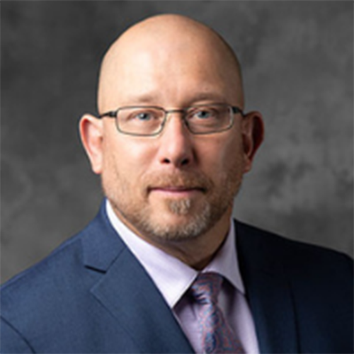 A man with glasses, a short beard, and a suit in front of a grey background