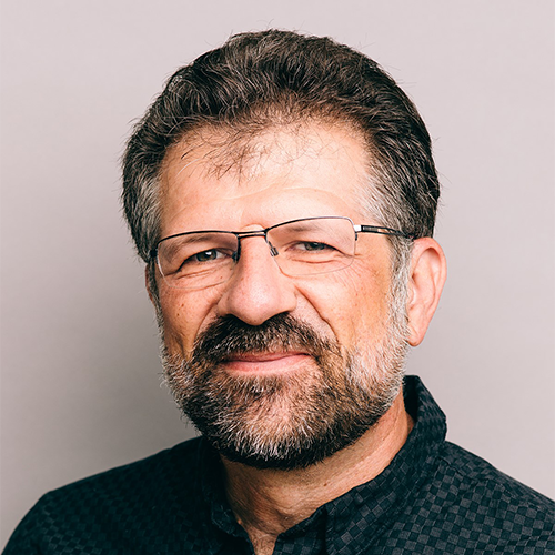 A man with short dark hair and a beard, wearing glasses and a dark collared shirt