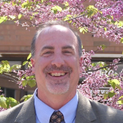 A man with grey hair and goatee, wearing a grey suit with blue shirt, standing in front of a tree with purple flowers