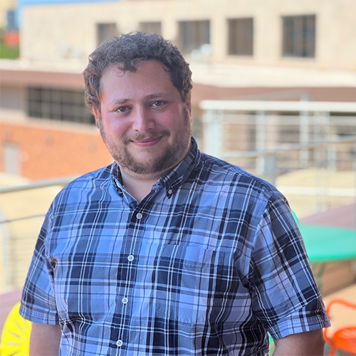 A man with short dark hair and a beard, wearing a blue plaid collared shirt, with a building in the background