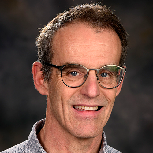 A man with short dark hair, a collared shirt, and glasses in front of a dark grey background