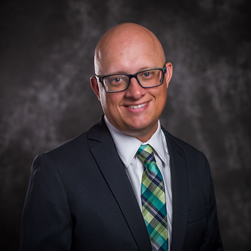 John Furcean, a man with glasses, wearing a dark suit jacket and green plaid tie. 