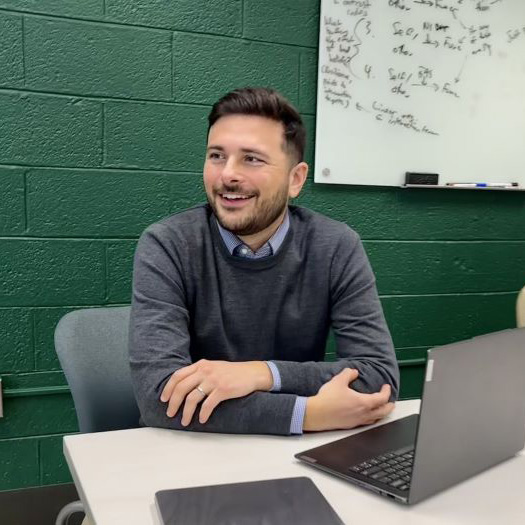 Henry Cowan near his computer and a whiteboard posing for a photo in the classroom