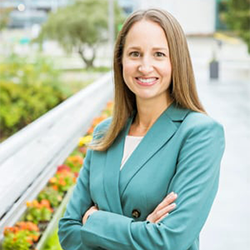 A woman with long light brown hair, wearing a teal blazer with her arms folded in front. She stands in front of a blurred background of trees and flowers.  