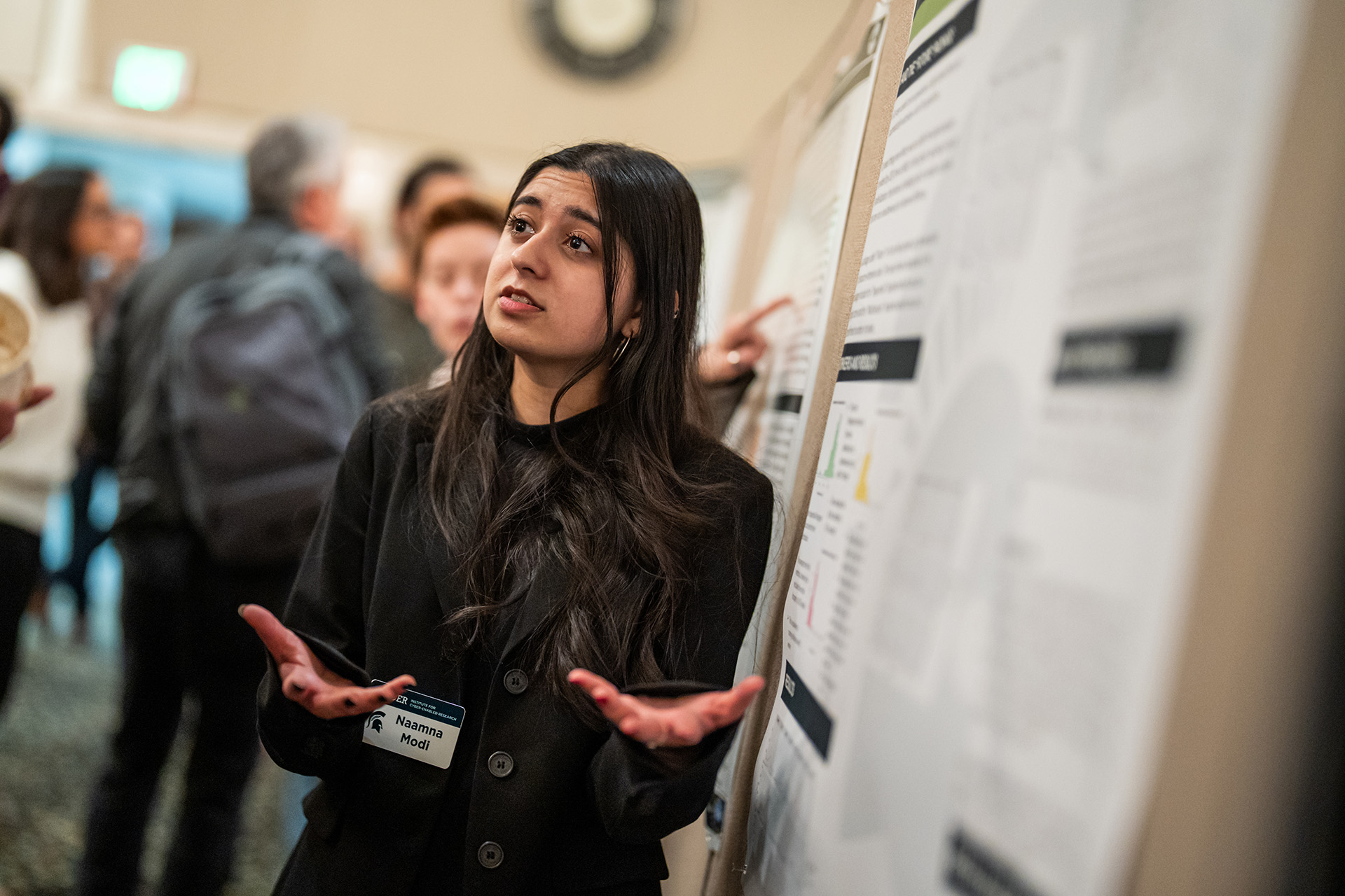 A student presents an academic poster in a crowded room.