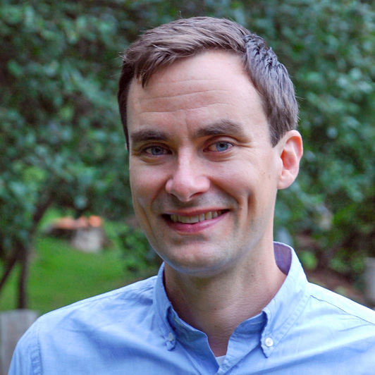 A man with short dark hair and blue shirt standing in a natural setting.