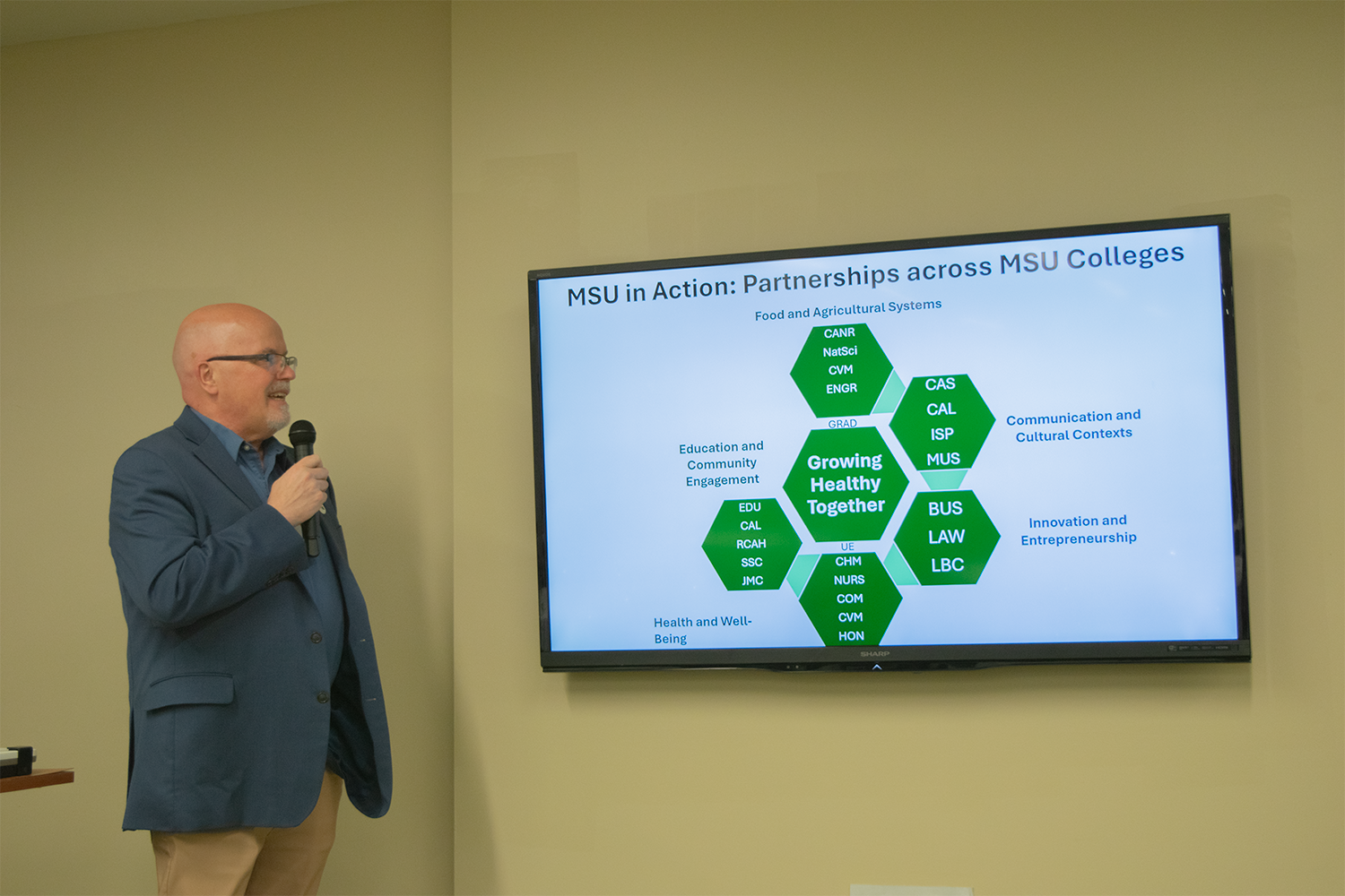 A man wearing a suit and glasses, holds a microphone and stands to the left of a TV on the wall with a slide with green hexagons and words on it.