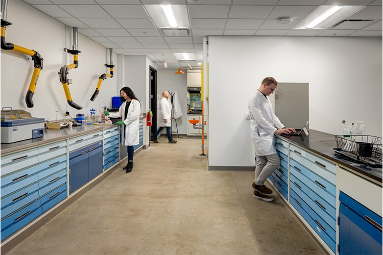 An image of a science lab, with two people standing at countertops, and one person walking in the back.