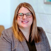 A woman with long reddish-brown hair, glasses, wearing a suit jacket in front of an ambiguous background.