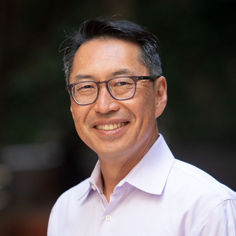 A man with dark hair, glasses, and light collared shirt in front of a dark background.