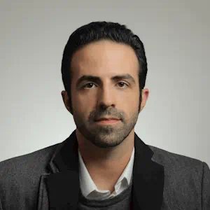 A man with short dark hair, white collared shirt and dark coat with thick lapel. 