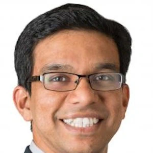 A smiling man with short dark hair, glasses, in front of a white background.