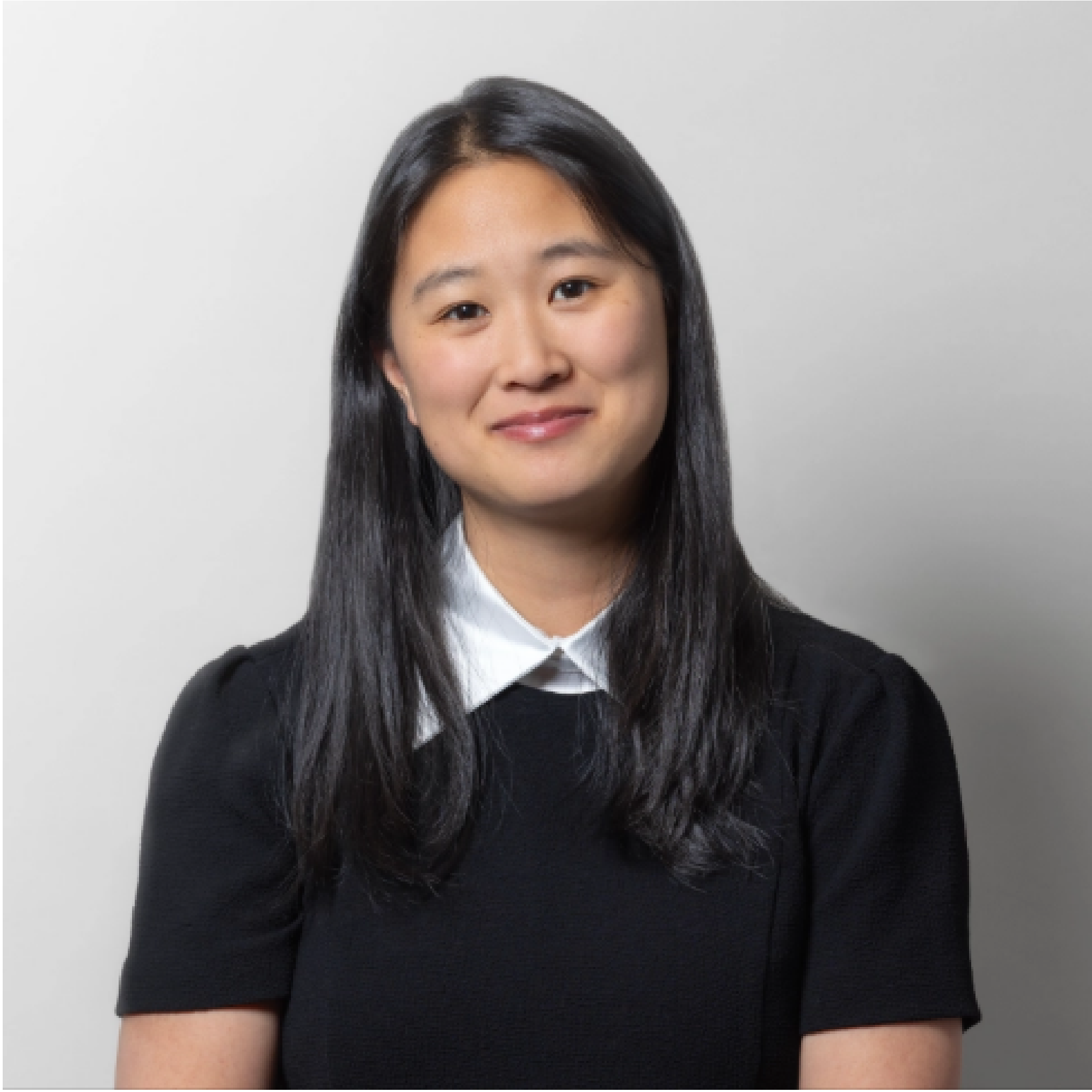 A woman with long black hair, wearing a black shirt with a white collar, in front of a light grey background.