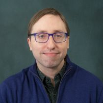 A man with short brown hair, glasses, wearing a dark blue jacket and dark shirt in front of a grey-green background.