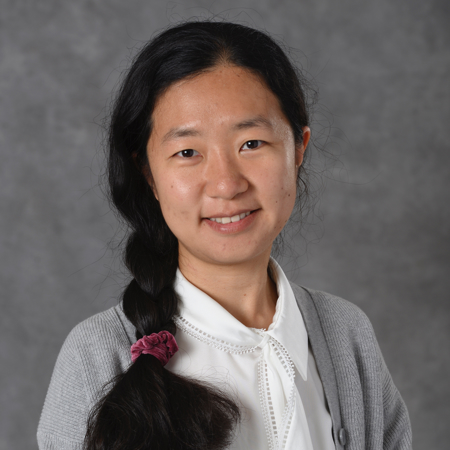 A woman with long black hair in a long braid along her right side. She wears a white blouse with a grey sweater in front of a grey background.
