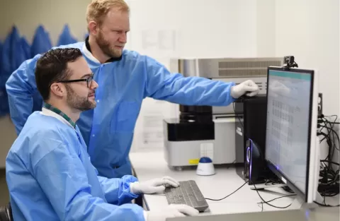Two men in blue scrubs in a lab looking at a computer
