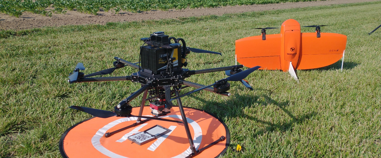 A large multirotor drone rests on an orange landing pad in a grassy field, with a bright orange fixed-wing drone positioned in the background.