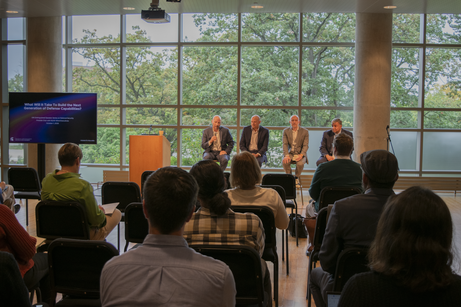 A room full of people sitting in chairs, listening to a panel of 4 people speaking in front of windows. 