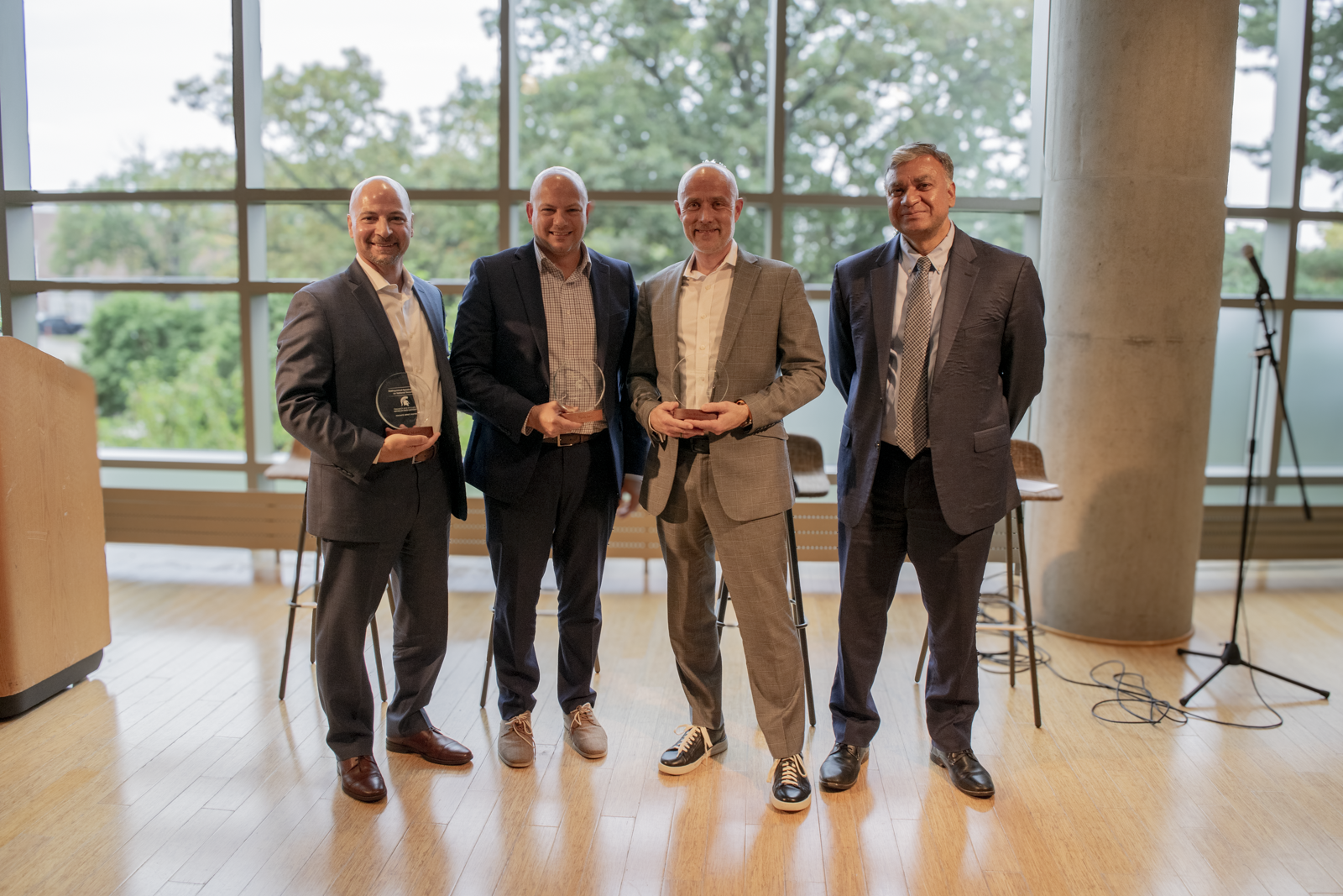 Four men in suits, standing in front of windows. Three of the men are holding an award. 