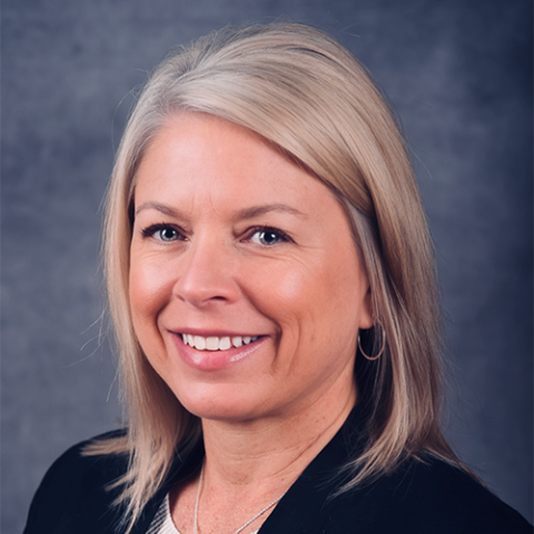 A woman with shoulder-length blonde hair, black jacket, in front of a grey background.