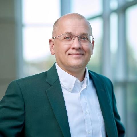 A man with very short hair, glasses, wearing a white collared shirt and dark green jacket