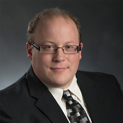 A man with short brown hair, glasses, wearing a tie, white shirt, and black suit jacket