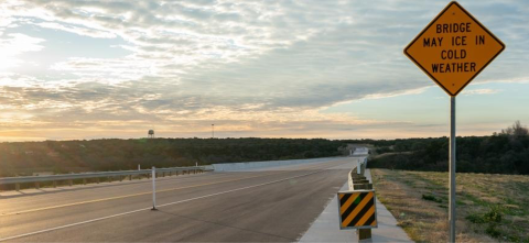 A warning sign that says “Bridge May Ice in Cold Weather” sits along a roadway with a bridge and sunset in the background.